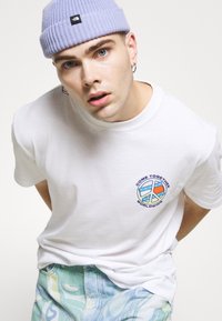 Hvid bomulds t-shirt med et cirkulært fredssymbol og teksten "KOM SAMMEN VERDEN OVER". Lavendel strikket hue og mønstrede lyse jeans.
