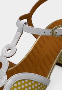 Sandale en cuir blanche avec un motif de découpes circulaires, une sangle accentuée d'or, un motif à pois jaunes et une semelle marron texturée.