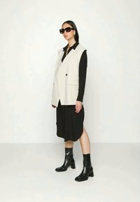 Colete bege sem mangas com dois bolsos frontais sobre uma camisa preta de manga longa e uma saia midi preta. Botas pretas de tornozelo e óculos de sol grandes.