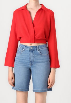 Blouse rouge courte à manches longues, avec un détail de nœud à l'avant. Associée à un short en jean taille haute bleu clair, retroussé au bas.