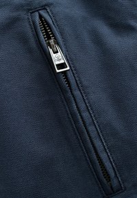 Fermeture éclair métallique partiellement ouverte sur un tissu bleu foncé texturé, montrant les dents et le détail du bord cousu.