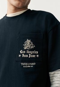 Mornarica pamučna majica s džepom na kojoj je bijela vezena ruža i tekst: "Los Angeles San Jose", "Tattoo Studio" i "441 West St."