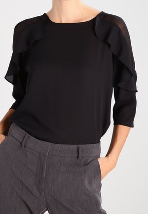 Blouse noire à manches à volants superposés, en tissu doux, avec un col rond ; associée à un pantalon gris ajusté avec des poches latérales.