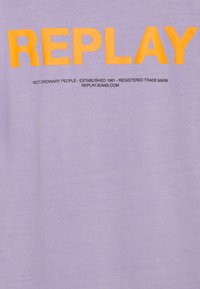 Fialové tričko s výrazným žlutým logem "REPLAY" a černým textem pod ním. Měkký materiál a volný střih.