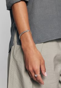 Srebrna warkoczowa bransoletka noszona na nadgarstku, zestawiona z pierścionkiem z prostokątnym kamieniem. Ręka jest umieszczona na szarym materiale.