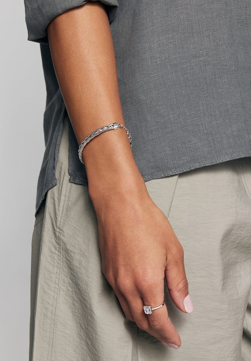 Srebrna warkoczowa bransoletka noszona na nadgarstku, zestawiona z pierścionkiem z prostokątnym kamieniem. Ręka jest umieszczona na szarym materiale.