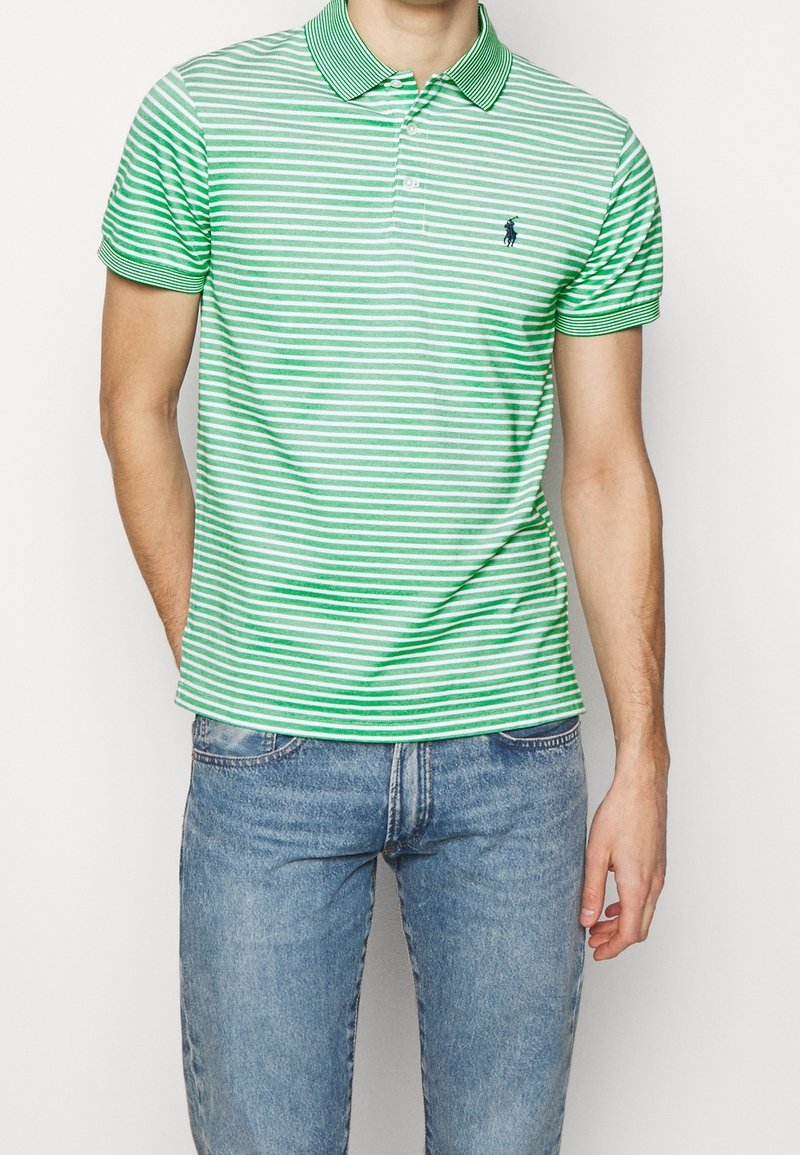 Homme portant un polo à rayures vertes et blanches avec un col et un jean bleu clair, debout avec une main dans la poche.