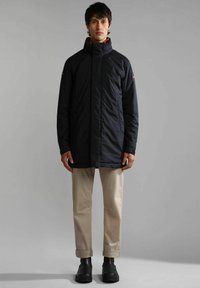 Giacca nera isolante con colletto alto, chiusura a zip e tasche laterali; indossata sopra pantaloni beige e stivali neri alla caviglia. Tessuto liscio.