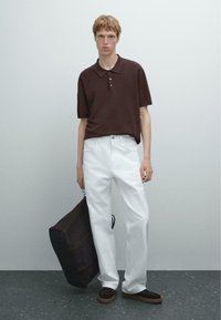 Polo shirt marrone a maniche corte, jeans bianchi a gamba dritta e sneaker marroni scuri. Borsa in pelle scura con tracolla tenuta nella mano sinistra.