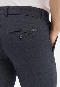 Donkerblauwe gestructureerde broeken met een achterzak voorzien van een knoopsluiting en een klein merklabel boven de zak.