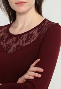 Bordeauxfarbendes Langarmoberteil mit floraler Spitze an der Schulternaht, rundem Ausschnitt und weichem, dehnbarem Stoff. Klassisches Design für vielseitige Tragemöglichkeiten.