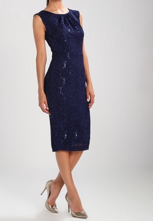 Robe midi sans manches en dentelle bleu marine avec des paillettes discrètes, portée avec des talons hauts métalliques à bout pointu sur un fond neutre.