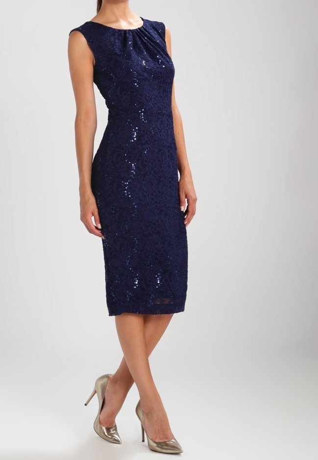 Robe midi sans manches en dentelle bleu marine avec des paillettes discrètes, portée avec des talons hauts métalliques à bout pointu sur un fond neutre.
