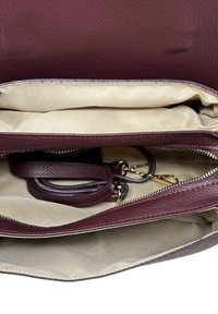 Bordowa skórzana torebka z dwoma przegrodami i kremową podszewką, wyposażona w kieszeń na suwak i odpinany pasek. Złote akcenty metalowe.