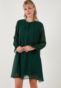 Zelena čipkasta haljina s dugim, prozirnim rukavima, okruglim izrezom i nabranim manžetama. Haljina pada iznad koljena i ima slobodan kroj.