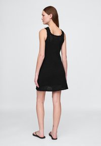 Crna kukičana haljina bez rukava, s valovitim trakama i proširenim rubom. Lagana pletena struktura omogućuje prozračnost.