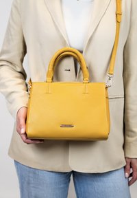 Geel faux leren handtas met dubbele handvatten, afneembare schouderriem en een gladde textuur. Voorzien van een zilverkleurig logo-accent.