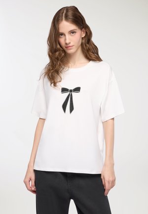 T-shirt bianco realizzato in morbido tessuto, con una grafica a forma di fiocco in paillettes nere sul davanti. Vestibilità ampia con maniche corte.