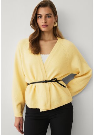 Kvinde iført en bleg gul cardigan med bælte over en hvid top, kombineret med sorte bukser, stående mod en ensfarvet væg.