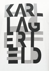 Μαύρα κεφαλαία γράμματα που σχηματίζουν το όνομα "KARL LAGERFELD", καλυμμένα με λεπτές οριζόντιες και κάθετες μαύρες γραμμές σε λευκό φόντο.