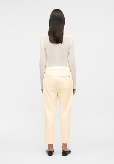 Γυναίκα με ίσια σκούρα μαλλιά φοράει μπεζ μπλούζα με μακριά μανίκια, ανοιχτόχρωμο κρεμ παντελόνι και μαύρα φλατ παπούτσια, στέκεται γυρισμένη προς τα πίσω σε απλό φόντο.