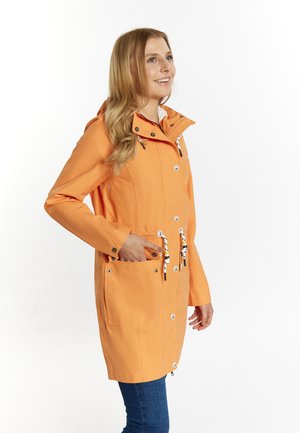 Vrouw glimlacht, draagt een lange oranje regenjas met witte koordjes, staat zijaanzicht met één hand in de zak.
