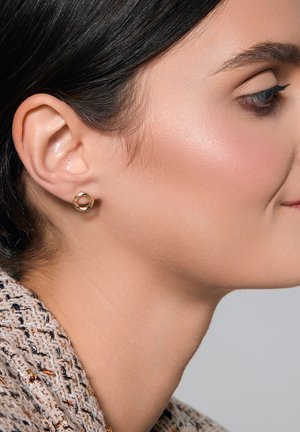 Złote kolczyki w kształcie obręczy o teksturowanym, okrągłym wzorze. Kolczyki noszone są na jasnej skórze, prezentując subtelny połysk.