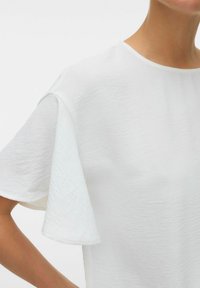 Haut court à manches courtes de couleur blanche en tissu texturé. Encolure ronde, avec des manches amples au design drapé et doux.