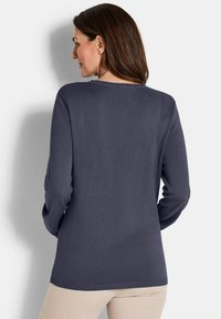 Frau mit braunen Haaren, die einen langärmligen dunkelgrauen Pullover und eine hellbeige Hose trägt, mit dem Rücken zur Kamera vor einem weißen Hintergrund.