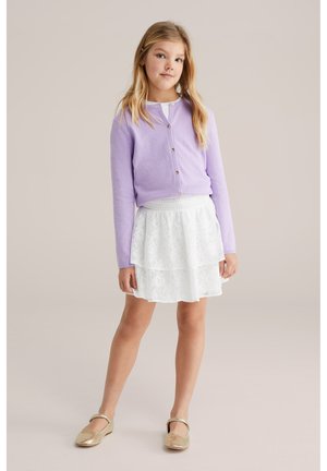 Mlada deklica stoji oblečena v sivkasto gumbasto jopico, belo čipkasto krilo in zlate čevlje Mary Jane, na enobarvnem ozadju.