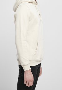 Sweat à capuche crème avec une poche kangourou, des cordons de serrage et des poignets côtelés. Confectionné dans un tissu doux, il présente une coupe décontractée et un design simple.