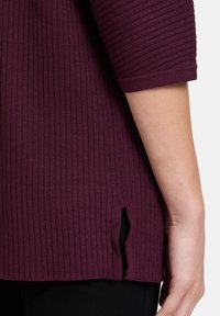 Pravá ruka vedle pasu, oblečená v tmavě fialovém krátkém rukávu s jemným bočním rozparkem, přes černé kalhoty.