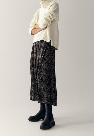 Vrouw met een crème coltrui, een geplooide midi-rok met slangenprint en zwarte chunky laarzen, staand met gekruiste armen.