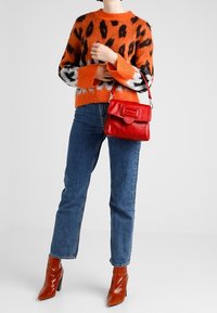 Sac à main en cuir rouge avec rabat et détails en métal, porté avec un pull orange et noir à motif et un jean bleu. Bottes marron à bout pointu.