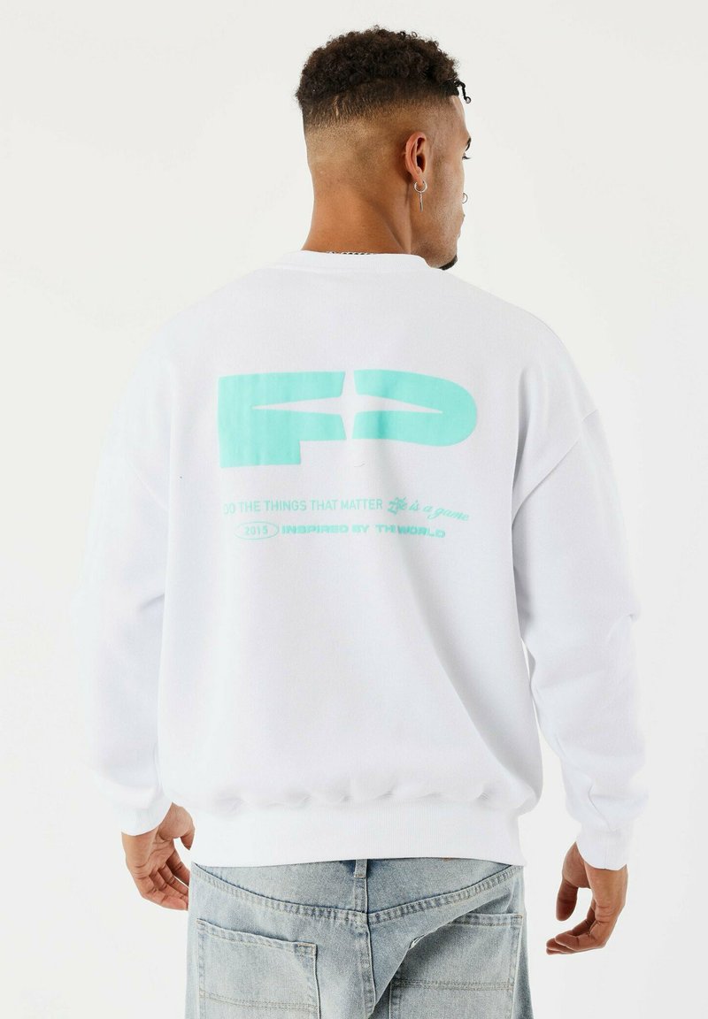 Hombre con cabello rizado corto que lleva puesta una sudadera blanca con texto y logo turquesa en la parte trasera, combinado con jeans de color azul claro.