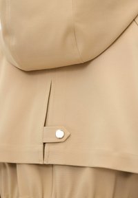 Beige vattentät jacka med texturerad tyg, huva och en synlig tryckknappsstängning på baksidan med sömnadsdetaljer.