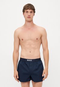 Șorturi de baie de un bleumarin închis, cu un talie ajustată, prevăzute cu logo-ul "BOSS." Materialul pare neted și ușor, având un detaliu cu nasture.