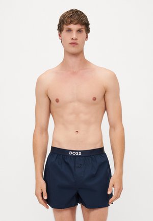 Dunkelmarineblaue Badehose mit einem enganliegenden Taillenbund, auf dem das Logo "BOSS" prangt. Der Stoff wirkt glatt und leicht und hat ein Knopfdetail.