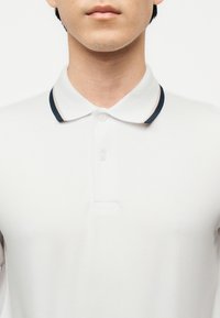 Άνδρας ντυμένος με λευκό πόλο πουκάμισο με γιακά που έχει λεπτές μαύρες και μπεζ ρίγες, εμφανιζόμενος από το λαιμό έως το πάνω μέρος του κορμού.