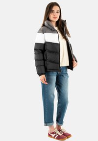 Giubbotto imbottito nero, grigio e bianco con zip frontale e cappuccio, abbinato a jeans larghi blu e sneakers bordeaux.