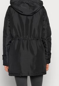 Doudoune noire avec taille cintrée, capuche et poignets à bouton-pression. Tissu lisse et brillant, longueur jusqu'à mi-cuisse.