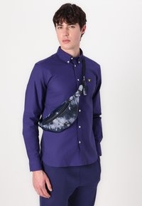 Camisa de manga larga morada con cierre de botones, llevada con una riñonera de teñido anudado azul marino que cuenta con un compartimento con cremallera y una correa ajustable.