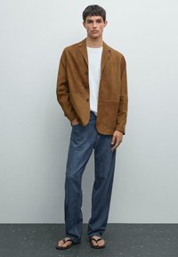 Brauner Wildleder-Blazer mit Reverskragen, drei Knöpfen und zwei Vordertaschen, kombiniert mit lockeren blauen Jeans und Flip-Flops.