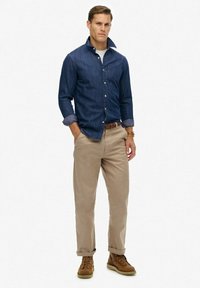 Donkerblauwe denim shirt met button-down kraag, beige broek en bruine laarzen. Het shirt heeft opgerolde mouwen en een borstzakje.