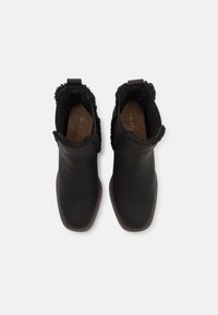 Para czarnych skórzanych botków za kostkę z futrzanym wykończeniem i brązowymi podeszwami, widocznych z góry na białym tle.