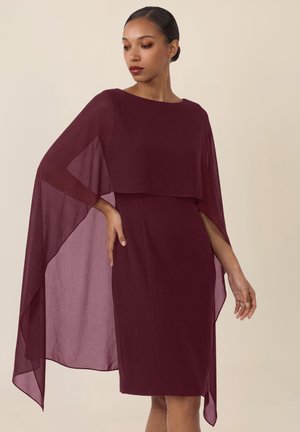 Frau trägt ein ärmelloses bordeauxfarbenes Kleid mit passendem transparentem Cape, steht mit einer Hand in der Hüfte vor einem schlichten Hintergrund.