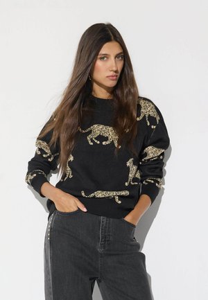 Joven mujer con pelo largo y castaño, vistiendo un suéter negro con estampado de leopardo beige y vaqueros oscuros de cintura alta con las manos en los bolsillos sobre fondo blanco.