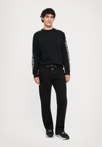 Melnā sporta jakā ar kontrastējošiem baltiem uzrakstiem uz piedurknēm, apvienota ar melnām džinsām. Modelis stāv pretī gaišai fona, valkājot melnus sporta apavus.