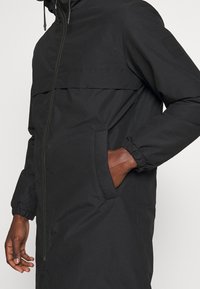 Homme portant une longue veste noire à capuche avec fermeture éclair à l'avant et poches latérales, main droite dans la poche sur un fond uni.