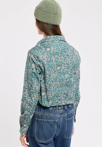 Langärmliges Shirt aus teal Baumwolle mit floralen und Paisley-Mustern; verfügt über einen Kragen und eine hintere Schulternaht. Kombiniert mit hochtaillierten Jeans.